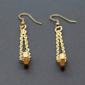 May include: A pair of gold-colored earrings. Each earring features a chain design with a small, geometric, gold-colored pendant at the bottom. The earrings have a hook closure. The background is a solid, dark gray color.