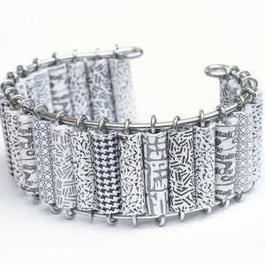 May include: A silver bracelet with a series of white paper tubes with black patterns. The tubes are connected by a silver chain.