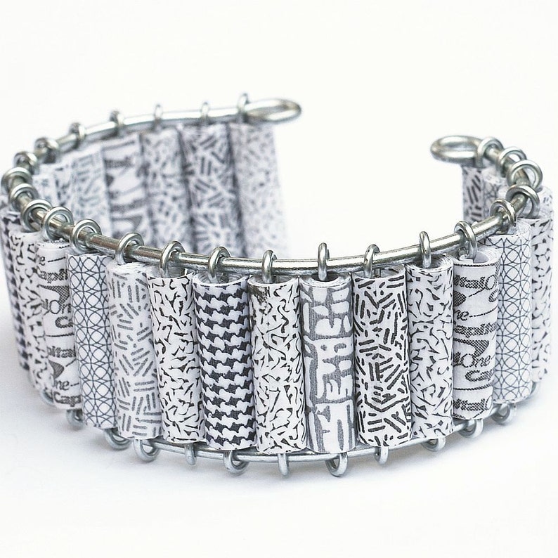 May include: A silver bracelet with a series of white paper tubes with black patterns. The tubes are connected by a silver chain.
