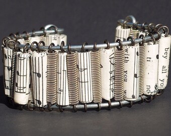 May include: A handmade bracelet featuring rolled paper segments printed with musical notes, connected by metal wire and springs. The bracelet has a unique, artistic design with a vintage aesthetic. The black and white color scheme gives it a classic look.