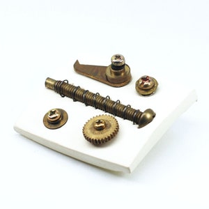 May include: A collection of brass gears, screws, and a small metal hook on a white surface. The items are arranged in a random pattern.