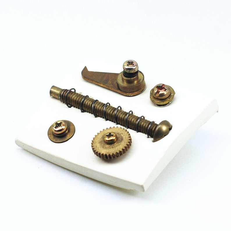 May include: A collection of brass gears, screws, and a small metal hook on a white surface. The items are arranged in a random pattern.