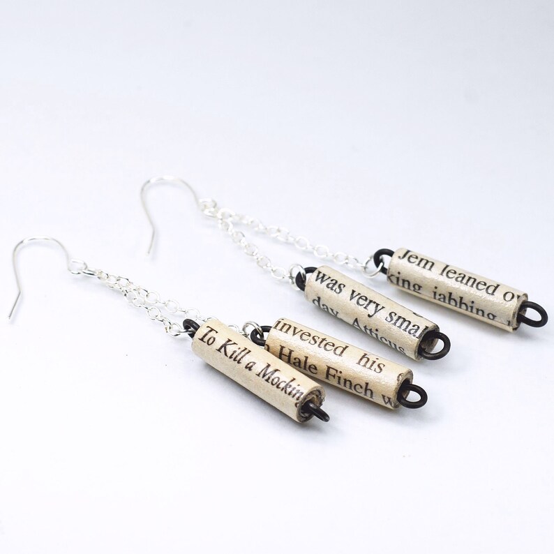 May include: Silver earrings with a chain and three small paper scroll charms. The charms are made from pages of the book "To Kill a Mockingbird" and feature text from the book.