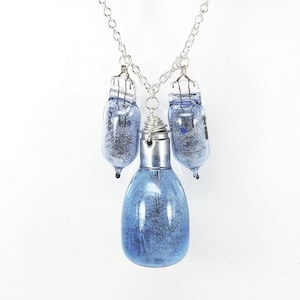Può includere: Una collana unica con tre ciondoli a forma di lampadina in vetro blu. La lampadina centrale è più grande, con un cappuccio in metallo argentato e un dettaglio a spirale di filo. Le lampadine sono sospese a una catena d'argento, un accessorio distintivo.