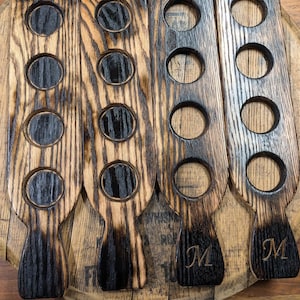 Puede incluir: Cuatro soportes de madera para vasos de chupito con un acabado negro quemado. Cada soporte tiene cinco recortes redondos y un asa. Los soportes est&aacute;n grabados con la letra "M".