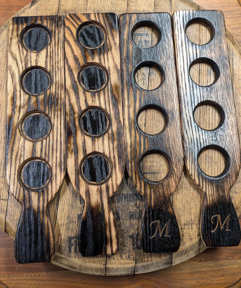 Puede incluir: Cuatro soportes de madera para vasos de chupito con un acabado negro quemado. Cada soporte tiene cinco recortes redondos y un asa. Los soportes est&aacute;n grabados con la letra "M".