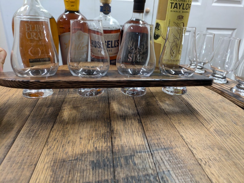 Puede incluir: Una bandeja de madera con seis vasos para degustar whisky. La bandeja est&aacute; hecha de madera reciclada y tiene un acabado marr&oacute;n oscuro. Los vasos son todos transparentes y tienen una forma redonda. Hay cinco botellas de whisky en el fondo, incluyendo Elijah Craig, Russell's Reserve y Colonel E.H. Taylor Jr. Single Barrel.