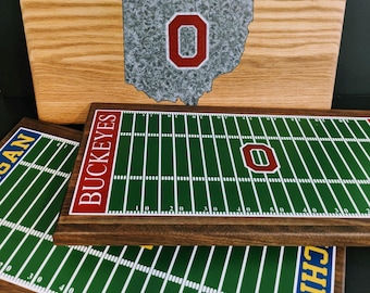 Puede incluir: Signos de madera con un contorno de Ohio con un logotipo "O" rojo. Debajo, signos rectangulares de madera con un diseño de campo de fútbol verde, líneas blancas y la palabra "Buckeyes" en rojo.