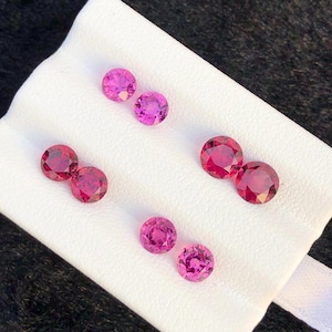 May include: Six round, faceted, vibrant pink and deep red gemstones arranged in pairs on a white display card. Each stone features a brilliant cut with multiple facets that reflect light, showcasing a circular shape and a polished, translucent finish.