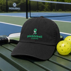 Op de afbeelding: Een zwarte baseballpet met een geborduurd groen pickleball-racket en bal-icoon boven de tekst "pickleball GRANDMA" in groene letters. De pet heeft een gebogen klep en een gestructureerde kroon met ventilatiegaatjes.