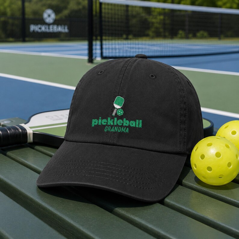 Op de afbeelding: Een zwarte baseballpet met een geborduurd groen pickleball-racket en bal-icoon boven de tekst "pickleball GRANDMA" in groene letters. De pet heeft een gebogen klep en een gestructureerde kroon met ventilatiegaatjes.