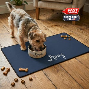 Może przedstawiać: Pies je z personalizowanej miski na granatowej macie do karmienia zwierząt. Miska jest beżowa z napisem "Lenny" w skrypcie. Mata ma również napis "Lenny" w skrypcie. Smakołyki dla psów w kształcie kości i śladów łap są rozrzucone.