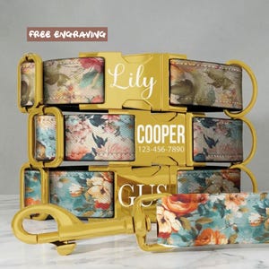 May include: Three pet collars and a leash featuring a floral pattern with orange, blue, and green tones. Each collar has a gold-colored metal buckle engraved with names Lily, COOPER, and GUS, plus a phone number on the middle buckle.