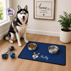 Peut inclure: Un tapis de nourriture pour animal de compagnie bleu marine rectangulaire avec un graphique blanc représentant une tête de husky et le nom Molly en écriture blanche. Le tapis porte deux bols en acier inoxydable sur un sol en bois.