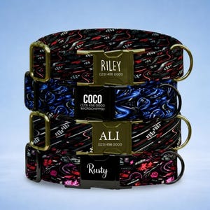 May include: Four dog collars stacked on top of each other. The collars have a black background with red, white, and blue designs. Each collar has a name tag with a name and phone number. The names are Riley, Coco, Ali, and Rusty.