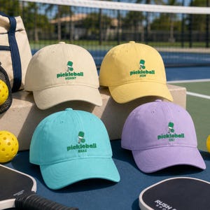 Op de afbeelding: Vier baseballpetten in beige, geel, turquoise en lavendel, elk voorzien van een geborduurd groen pickleball-racket en bal-icoon met de tekst pickleball gevolgd door MOMMY, MOM, NANA of GIGI gecentreerd op de voorzijde.