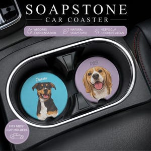 May include: Two round soapstone car coasters placed in a vehicle cup holder. The left coaster is light blue with a black, tan, and white dog illustration and the name Dakota. The right coaster is lavender with a beagle illustration and the name Bailey.