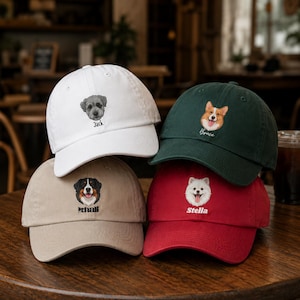 Può includere: Quattro cappellini da baseball bianchi, verdi, beige e rossi. Ogni cappellino presenta una razza di cane diversa ricamata sul davanti, con il nome del cane sottostante. I cappellini sono esposti su una superficie di legno.