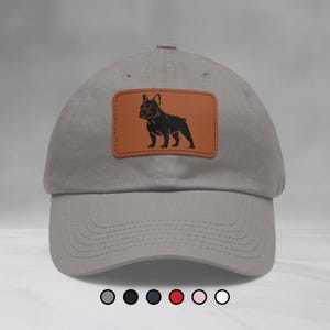 Peut inclure: Casquette de baseball grise avec un patch en cuir marron représentant une silhouette noire d'un bouledogue français. La casquette a une visière incurvée et un design cousu. Le fond est gris neutre.