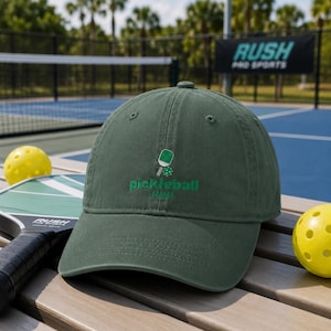 Op de afbeelding: Een donkergroene baseballpet met een geborduurde afbeelding van een pickleball-racket en een bal, met de tekst "pickleball MAMA" in groen garen op het voorpaneel gestikt. De pet heeft een gebogen klep en een gestructureerde kroon.