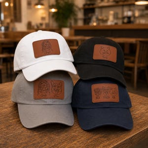 May include: Four baseball caps in white, black, gray, and navy blue are stacked on a wooden surface. Each cap has a brown leather patch with a pet illustration and name. The caps feature a curved brim and adjustable strap.