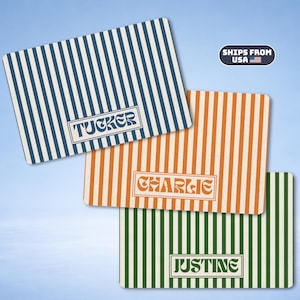 Peut inclure: Trois tapis rectangulaires à rayures verticales bleues, orange et vertes. Chaque tapis présente un cadre rectangulaire central contenant un nom dans une police stylisée : TUCKER sur le tapis bleu, CHARLIE sur l'orange et JUSTINE sur le vert.