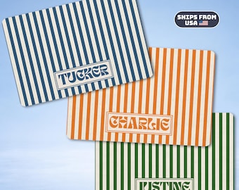 May include: Three rectangular mats featuring vertical stripes in blue, orange, and green. Each mat displays a centered rectangular frame containing a name in a stylized font: TUCKER on the blue mat, CHARLIE on the orange mat, and JUSTINE on the green mat.