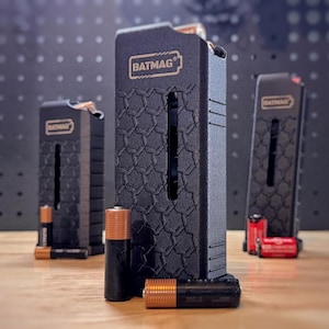 May include: Three black rectangular battery storage cases with the text BATMAG printed on the front. Each case features a textured hexagonal pattern and a vertical slot. Several AA and AAA batteries are positioned in front of and inside the cases.