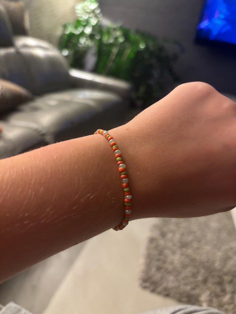 K&ouml;nnte beinhalten: Ein Armband aus kleinen, bunten Perlen in Orange-, Gr&uuml;n- und Wei&szlig;t&ouml;nen.