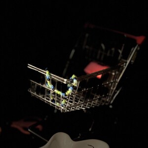 K&ouml;nnte beinhalten: Ein kleiner, silberner Einkaufswagen mit einer blauen und gr&uuml;nen Perlenkette darin.