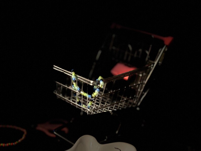 K&ouml;nnte beinhalten: Ein kleiner, silberner Einkaufswagen mit einer blauen und gr&uuml;nen Perlenkette darin.