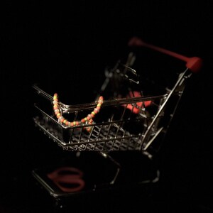 K&ouml;nnte beinhalten: Ein kleiner, silberner Einkaufswagen mit einer bunten Perlenkette darin. Der Wagen steht auf einem schwarzen Hintergrund.