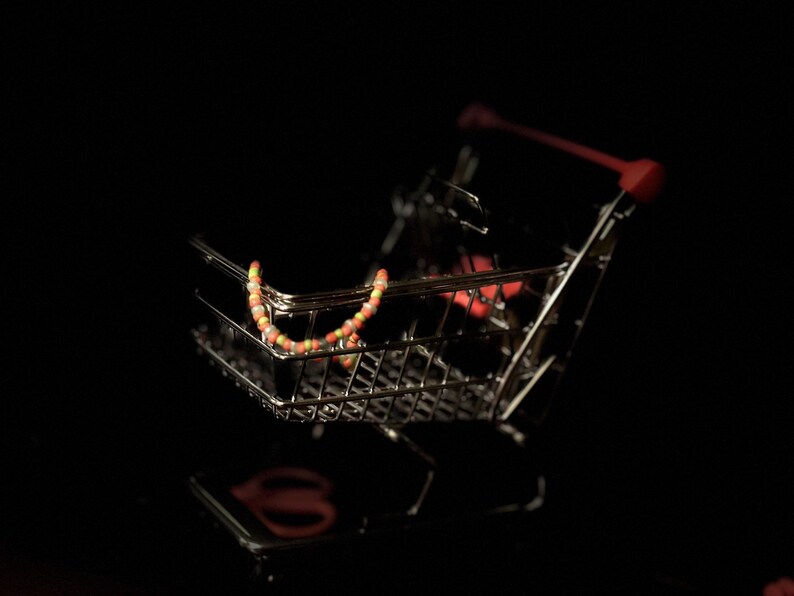 K&ouml;nnte beinhalten: Ein kleiner, silberner Einkaufswagen mit einer bunten Perlenkette darin. Der Wagen steht auf einem schwarzen Hintergrund.