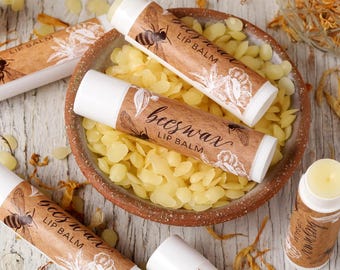 May include: A close-up of a bowl filled with yellow beeswax flakes. Several tubes of beeswax lip balm are arranged around the bowl. The lip balm tubes have brown paper labels with the text "beeswax lip balm" printed on them.