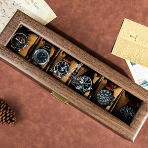 May include: A wooden watch box with a glass top, displaying six watches with different styles and colors. The box has a brass latch and is sitting on a brown surface.