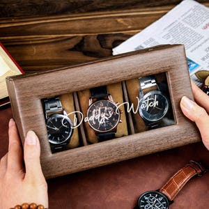 Può includere: Un porta orologi in legno con un coperchio trasparente, che mostra tre orologi. La scatola è incisa con le parole "Daddy's Watch".