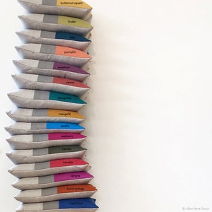 May include: A stack of 15 decorative throw pillows with different colored fabric panels. The pillows are arranged vertically with the colored panels facing the viewer. The colors of the panels are butternut squash, linden, lake, pumpkin, amethyst, sienna, biscay bay, marigold, pacific, raspberry, cypress, marsala, sangria, burnt orange, and royal blue.