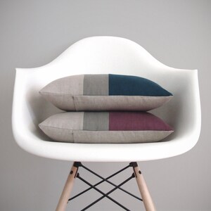 May include: Two decorative throw pillows with geometric patterns in shades of gray, teal, and burgundy. The pillows are placed on a white chair with a wooden base.