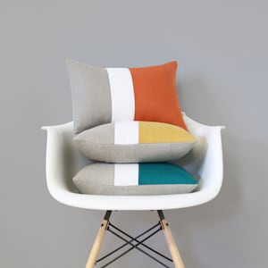 May include: Three decorative throw pillows stacked on a white chair. The pillows are made of a linen fabric and feature a striped design in shades of gray, white, orange, yellow, and teal.