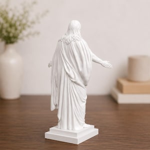 Pode incluir: Est&aacute;tua branca de uma figura com cabelo comprido e uma t&uacute;nica drapeada, com os bra&ccedil;os estendidos. A escultura est&aacute; sobre uma base retangular branca, sobre um fundo neutro. A est&aacute;tua tem cerca de 30 cm de altura.