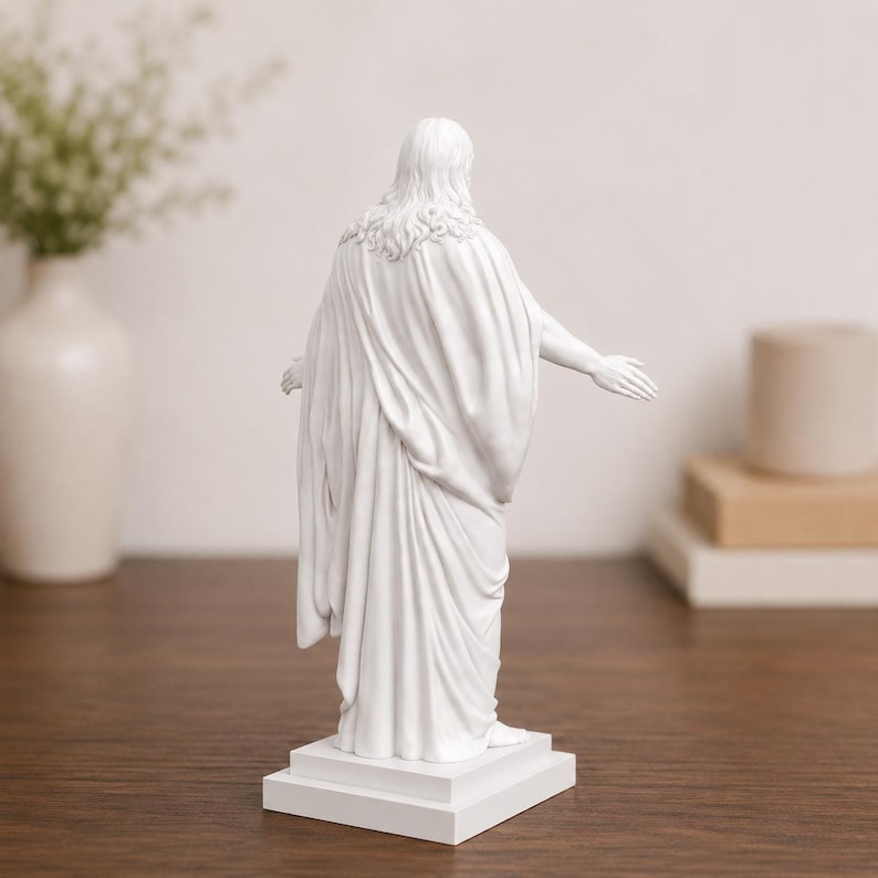 Pode incluir: Est&aacute;tua branca de uma figura com cabelo comprido e uma t&uacute;nica drapeada, com os bra&ccedil;os estendidos. A escultura est&aacute; sobre uma base retangular branca, sobre um fundo neutro. A est&aacute;tua tem cerca de 30 cm de altura.
