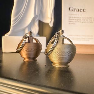 Könnte beinhalten: Zwei goldfarbene Schlüsselanhänger aus Metall in Form kleiner Urnen mit aufwendigen Designs. Jeder Schlüsselanhänger hat einen silbernen Ring.