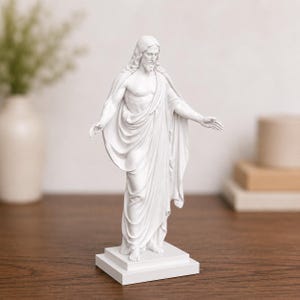 Pode incluir: Est&aacute;tua branca de uma figura com cabelo comprido e barba, vestida com vestes drapeadas, em p&eacute; sobre uma base quadrada. Os bra&ccedil;os da figura est&atilde;o estendidos. A est&aacute;tua est&aacute; sobre uma superf&iacute;cie de madeira.