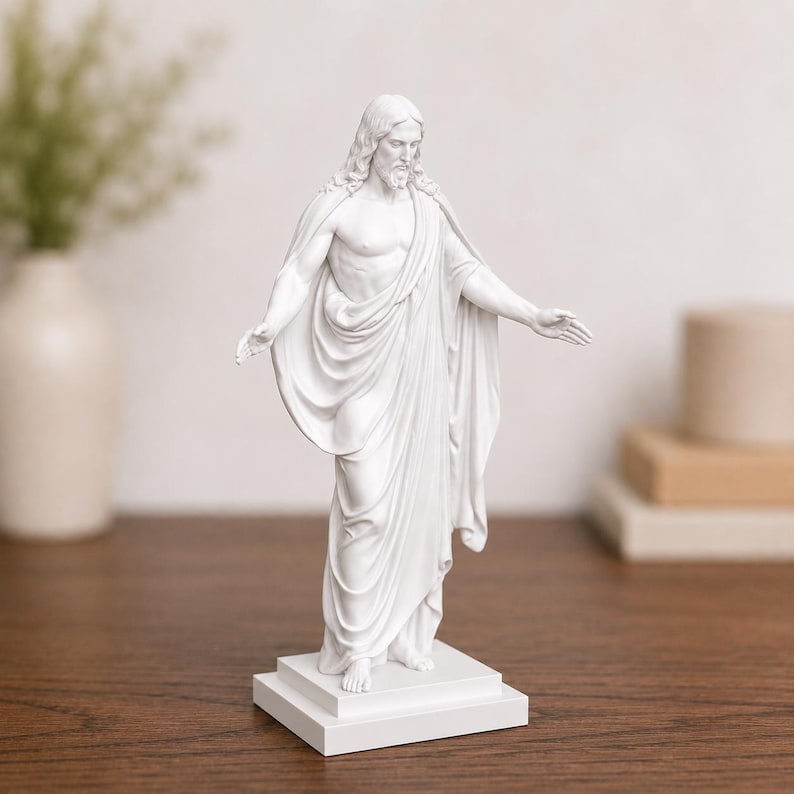 Pode incluir: Est&aacute;tua branca de uma figura com cabelo comprido e barba, vestida com vestes drapeadas, em p&eacute; sobre uma base quadrada. Os bra&ccedil;os da figura est&atilde;o estendidos. A est&aacute;tua est&aacute; sobre uma superf&iacute;cie de madeira.