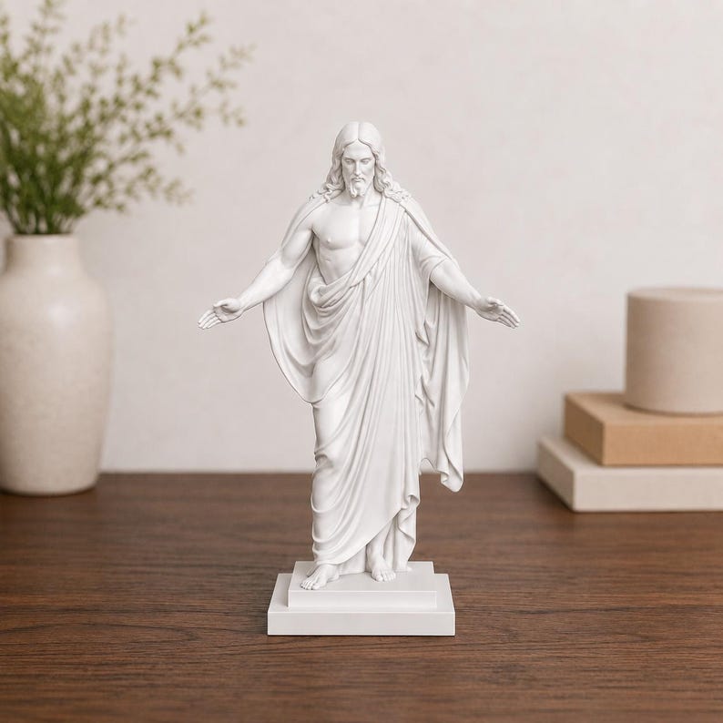 Pode incluir: Uma est&aacute;tua branca de uma figura com os bra&ccedil;os estendidos, envolta em vestes esvoa&ccedil;antes. A est&aacute;tua est&aacute; sobre uma base retangular e est&aacute; sobre uma superf&iacute;cie de madeira. A figura tem cabelo comprido e barba. Um vaso com vegeta&ccedil;&atilde;o est&aacute; no fundo.