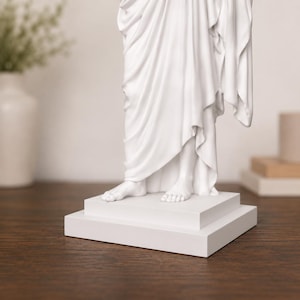 Pode incluir: Uma est&aacute;tua branca de uma figura drapeada, com p&eacute;s vis&iacute;veis e tecido esvoa&ccedil;ante. A escultura est&aacute; sobre uma base retangular em camadas. A est&aacute;tua est&aacute; sobre um fundo neutro, com uma superf&iacute;cie de madeira em primeiro plano.