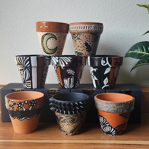 May include: A collection of eight terracotta flower pots, each hand-painted with unique designs. The pots feature various patterns, including moons, spiders, and floral motifs, in colors like orange, black, and white. Some pots have textured rims.