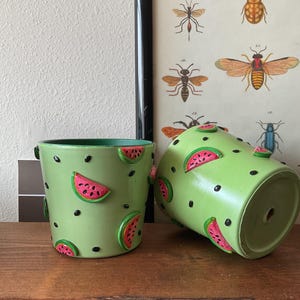 Puede incluir: Dos macetas de cerámica verde con un patrón de sandía. Las macetas están decoradas con semillas negras y rodajas de sandía rosadas.