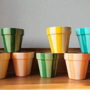 Puede incluir: Una colección de diez pequeñas macetas pintadas a mano. Cada maceta está pintada con rayas verticales en varios colores, incluyendo rojo, verde, amarillo, naranja y turquesa. Las macetas están dispuestas sobre una superficie de madera.