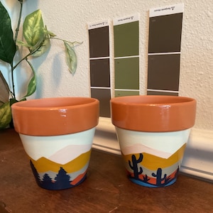 May include: Two hand-painted terracotta flower pots. Each pot features a landscape design with a terracotta rim, white upper section, and a colorful painted scene of mountains, trees, and cacti. The pots sit on a wooden surface.